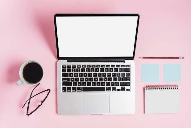 Een bovenaanzicht van koffiekopje; bril; laptop; potlood; zelfklevend blocnote en spiraalvormige blocnote tegen roze achtergrond
