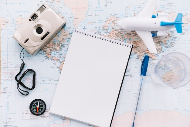 Een bovenaanzicht van miniatuur wit vliegtuig; spiraal lege blocnote; pen; camera en kompas op de kaart