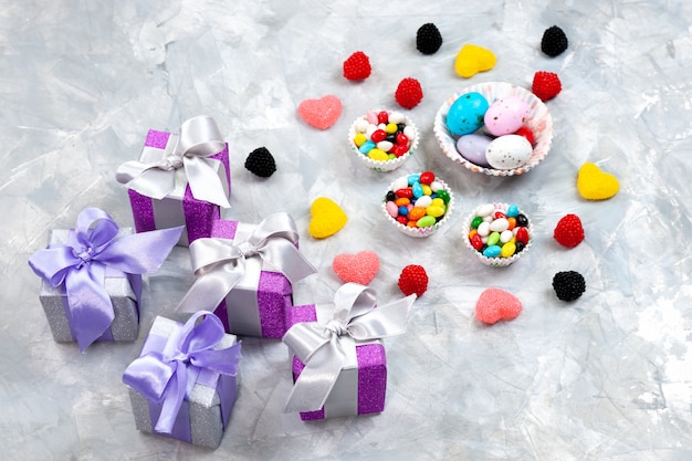Een bovenaanzicht veelkleurige snoepjes in kleine bordjes samen met hartvormige marmelades en paarse geschenkdozen op de grijze achtergrond verjaardag suiker viering regenboog