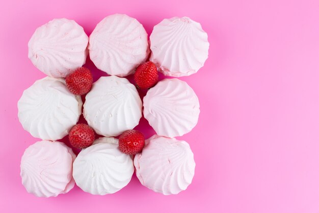 Een bovenaanzicht witte schuimgebakjes met verse rode aardbeien bekleed op roze bureau, koekjes zoetwaren zoete suikerkleur