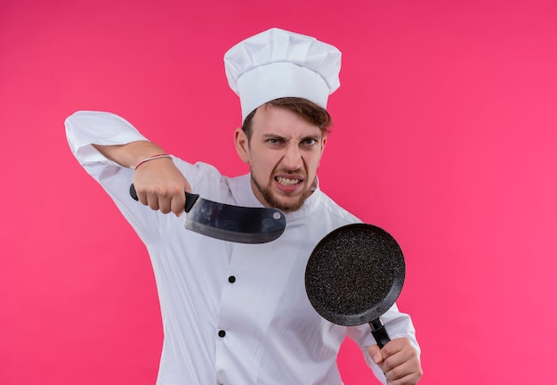 Een boze jonge, bebaarde chef-kokmens in wit uniform die vleesmes met koekenpan houdt terwijl hij op een roze muur kijkt