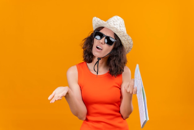 Een boze jonge vrouw met kort haar in een oranje overhemd dat zonnehoed en zonnebril draagt die kaartopenende handen houdt