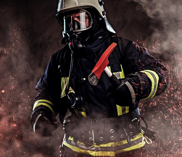 Een brandweerman gekleed in een uniform en een zuurstofmasker houdt een rode bijl vast die in vuurvonken staat en rook op een donkere achtergrond.