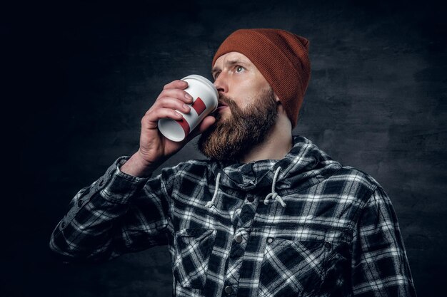 Een brutale bebaarde man gekleed in een hoed en fleece shirt, drinkt koffie uit een papieren glas.