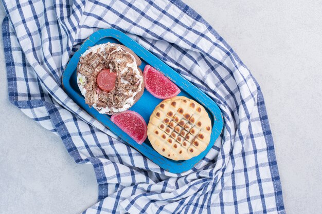 Een cake, een koekje en marmelades op een schaal op een handdoek op marmeren tafel.