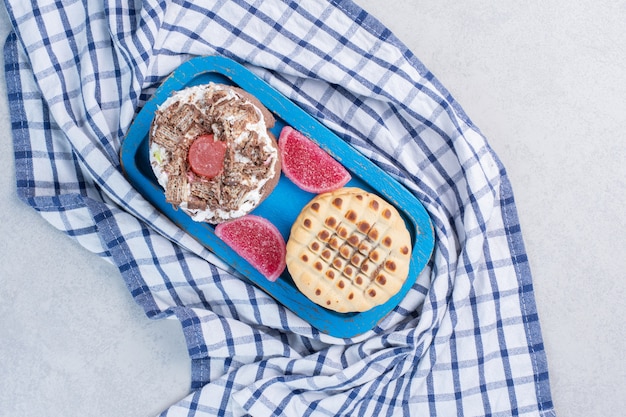 Een cake, een koekje en marmelades op een schaal op een handdoek op marmeren tafel.