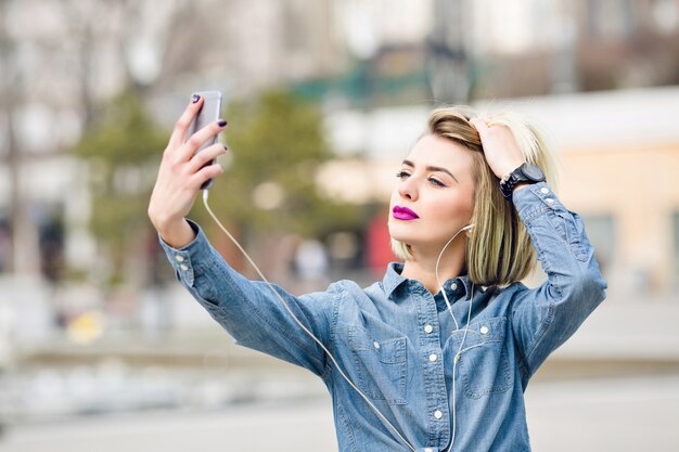 Een close-upportret van een dromerig blond meisje met felroze lippen die een selfie op een smartphone nemen