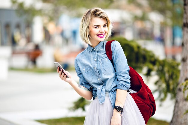 Een close-upportret van een staand lachend meisje met kort blond haar en felroze lippen met een smartphone