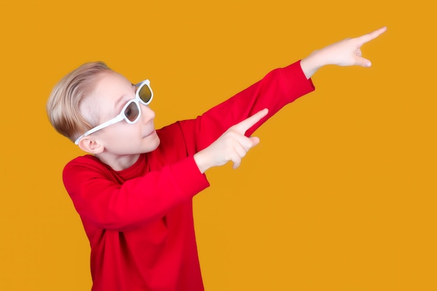 Een cool kind op een gele achtergrond en in een 3d-bril voor kinderen wijst met zijn handen de richting naar de zijkant