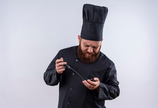 Een denkende bebaarde chef-kok in zwart uniform die zwarte pollepel met twee handen vasthoudt en ernaar kijkt op een witte muur