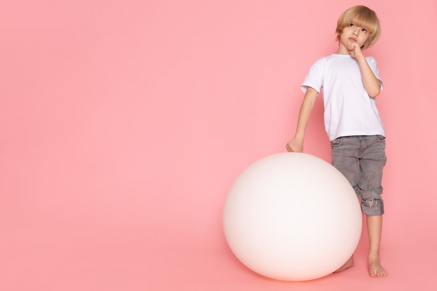 Een denkende jongen die van het vooraanzichtblonde in witte t-shirt met witte bal op het roze bureau legt