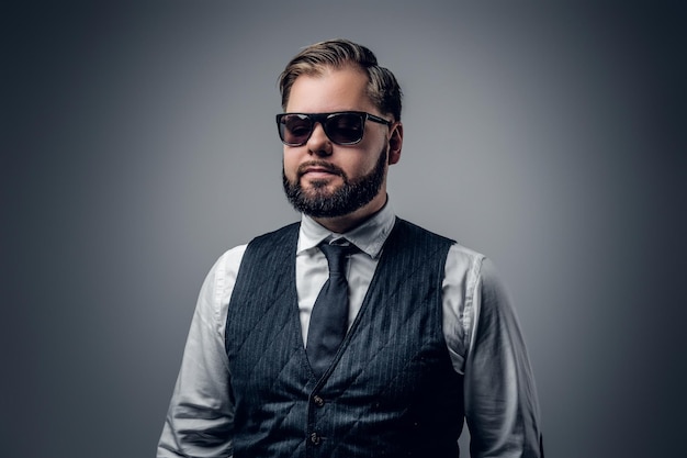 Een elegante bebaarde zakenman draagt een zonnebril en vest op een grijze achtergrond.