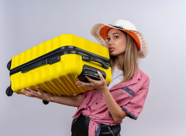 Een ernstige jonge vrouw die een rood overhemd en een zonnehoed draagt die gele koffer houdt terwijl zij op een witte muur kijkt