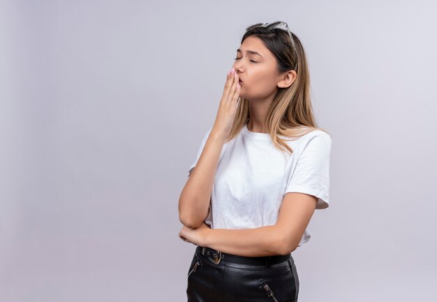 Een ernstige vrij jonge vrouw in wit t-shirt die zonnebril op haar hoofd draagt die hand op mond op een witte muur houdt