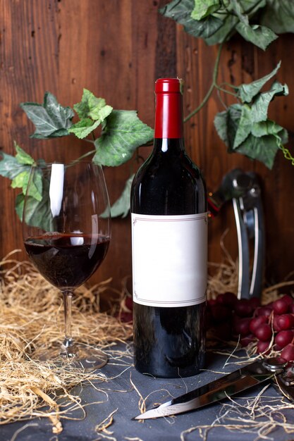 Een fles rode wijn van het vooraanzicht rode wijn samen met rode bessen en groene bladeren die op de grijze drank van de bureaualcoholwijnmakerij worden geïsoleerd