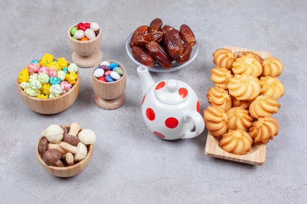 Een geassorteerde set koekjes, dadels, snoepjes en chocoladepaddestoelen naast een kleine theepot op marmeren oppervlak.