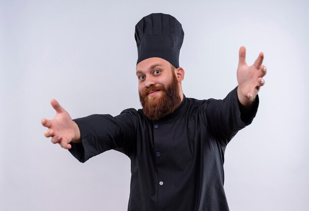 Een gelukkig bebaarde chef-kok in zwart uniform die zijn armen opent voor een knuffel terwijl hij naar de camera kijkt op een witte muur