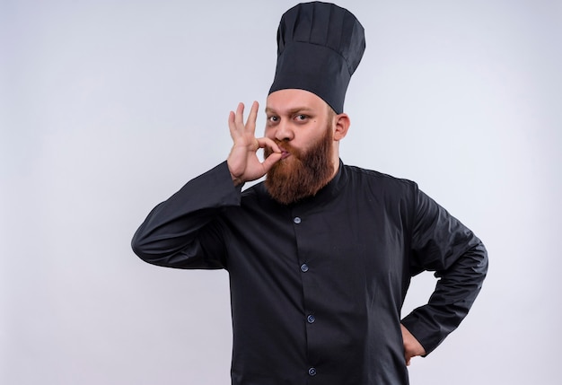 Een gelukkige bebaarde chef-kokmens in zwart uniform die ok gebaar toont terwijl hij naar de camera op een witte muur kijkt