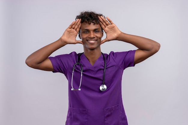 Een gelukkige jonge knappe donkere arts met krullend haar draagt violet uniform met een stethoscoop hand in hand op het hoofd