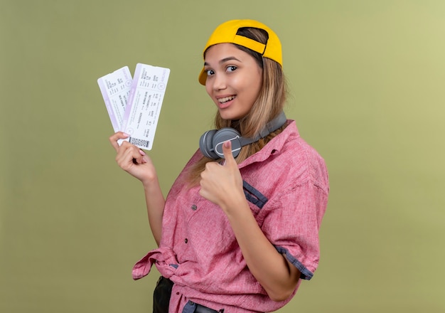 Een gelukkige jonge vrouw die een rood overhemd en een gele honkbalhoed draagt die duimen toont terwijl zij vliegtickets op een groene muur houdt