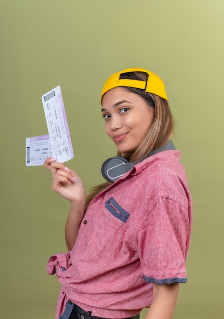 Gratis foto een gelukkige jonge vrouw die een rood overhemd en een gele honkbalhoed draagt die vliegtickets toont terwijl zij op een groene muur kijkt