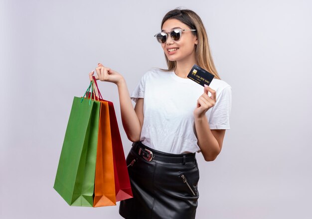 Een gelukkige jonge vrouw in wit t-shirt die zonnebril draagt die creditcard toont terwijl zij boodschappentassen op een witte muur houdt