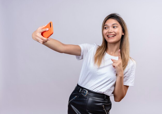 Een gelukkige jonge vrouw in wit t-shirt met koptelefoon die een selfie met smartphone neemt en met wijsvinger op een witte muur wijst