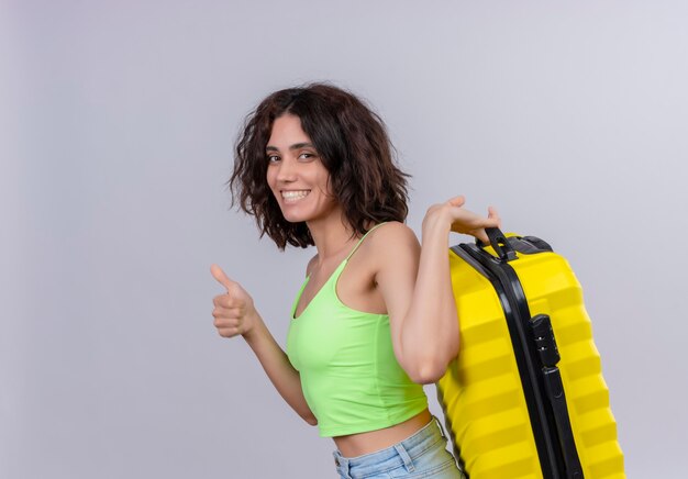 Een gelukkige jonge vrouw met kort haar in een groene crop top glimlachend en met gele koffer op een witte achtergrond