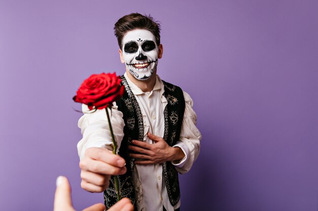 Een gelukkige kerel met een vrolijke blik is dankbaar en geeft roos aan zijn geliefde persoon. Binnenportret van de mens met halloween-make-up.