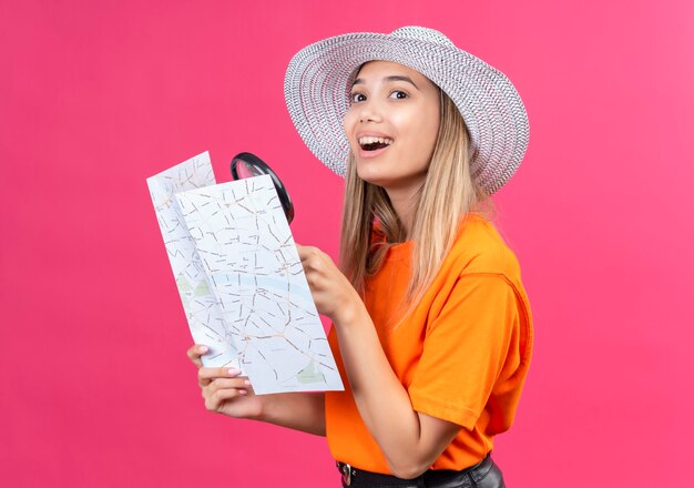 Een gelukkige mooie jonge vrouw in een oranje t-shirt met zonnehoed die een kaart met vergrootglas vasthoudt terwijl ze opzij kijkt op een roze muur