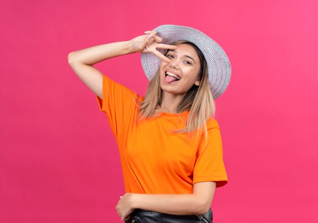 Een gelukkige mooie jonge vrouw in een oranje t-shirt met zonnehoed die haar tong met twee handengebaar toont terwijl zij op een roze muur kijkt