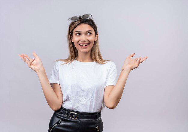 Een gelukkige mooie jonge vrouw in een wit t-shirt met een zonnebril op haar hoofd opent een groetgebaar in terwijl ze op een witte muur kijkt