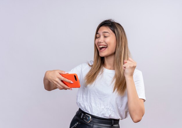 Een gelukkige mooie jonge vrouw in wit t-shirt die gebalde vuist opheft terwijl ze mobiele telefoon met gesloten ogen op een witte muur houdt