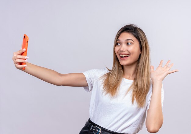 Een gelukkige mooie jonge vrouw in wit t-shirt praten over videogesprek met smartphone en zwaaiende hand hallo gebaar maken op een witte muur