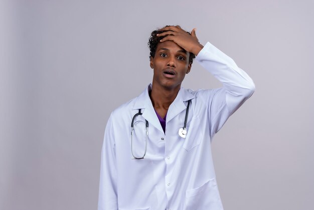 Een geschokte jonge knappe donkere arts met krullend haar, gekleed in een witte jas met een stethoscoop die de handpalm op het voorhoofd houdt
