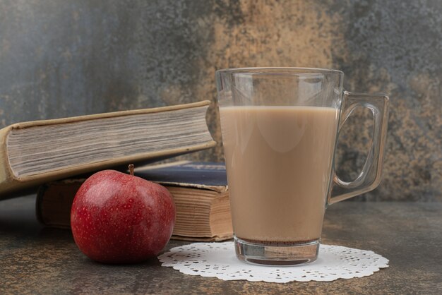 Een glas hete koffie met één rode appel en boeken over marmeren muur.