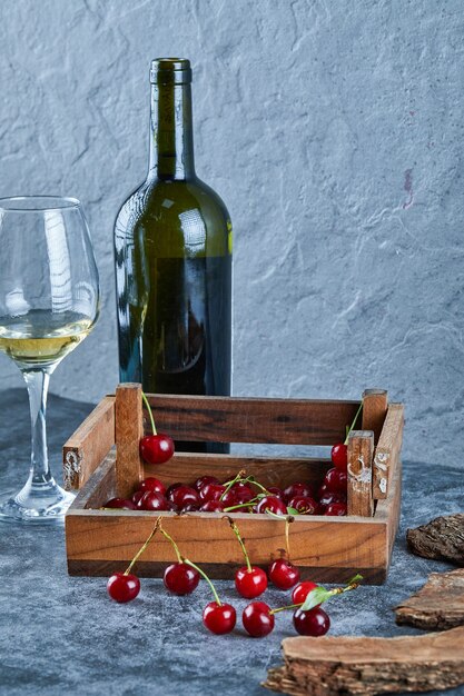 Een glas witte wijn, een fles en een houten kist met kersen op blauwe ondergrond