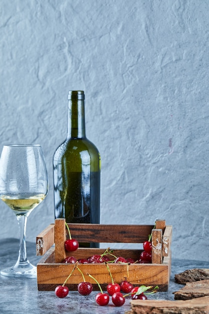 Een glas witte wijn, een fles en een houten kist met kersen op blauwe ondergrond