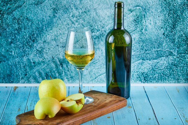 Een glas witte wijn en een fles met appelschijfjes op blauwe muur.