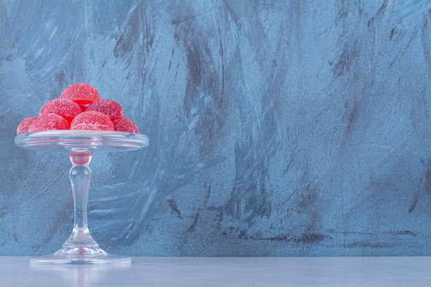 Een glazen plaat vol rode suikerachtige fruitgelei snoepjes op grijze tafel.