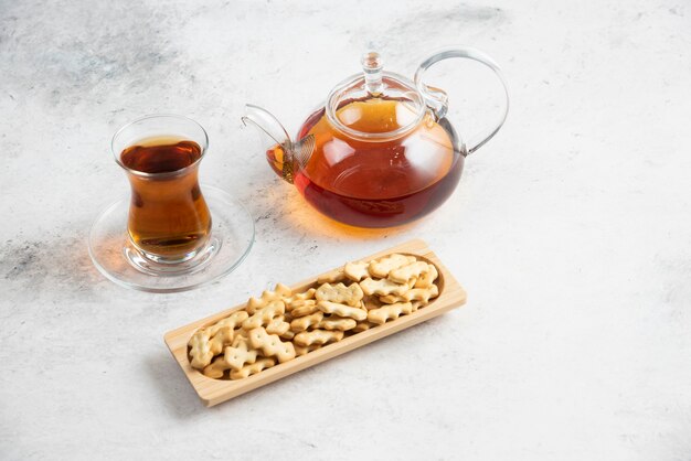 Een glazen theepot met een kopje thee en een houten plank vol crackers.