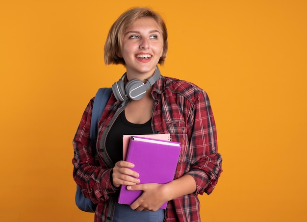 Een glimlachend jong slavisch studentenmeisje met een koptelefoon om de nek die een rugzak draagt, houdt een boek en een notitieboekje vast en kijkt naar de zijkant
