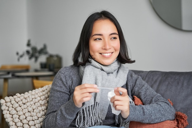 Een glimlachende aziatische vrouw die een thermometer vasthoudt en er tevreden uitziet, voelt zich beter nadat ze verkouden is geworden en fev kwijt is