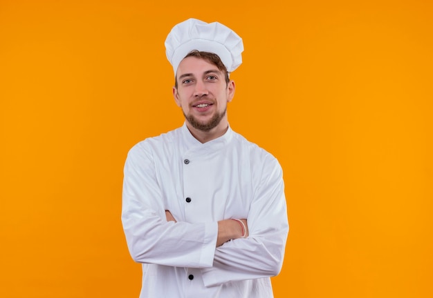 Een glimlachende jonge bebaarde chef-kok man in wit uniform hand in hand gevouwen terwijl hij op een oranje muur keek