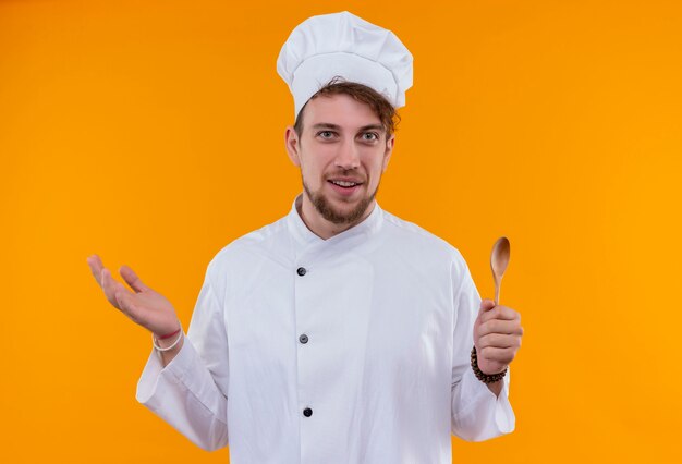 Een glimlachende jonge, bebaarde chef-kokmens in wit uniform houdt houten lepel vast terwijl hij op een oranje muur kijkt