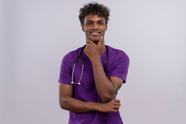 Een glimlachende jonge knappe donkere arts met krullend haar die violet uniform met een stethoscoop draagt die hand op kin houdt