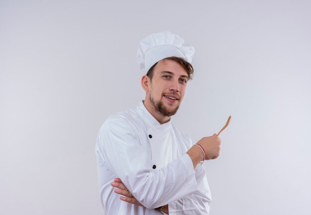 Een glimlachende knappe jonge bebaarde chef-kok man in wit fornuis uniform en hoed met houten lepel terwijl het kijken op een witte muur