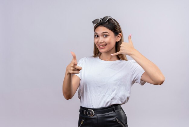 Een glimlachende mooie jonge vrouw in een wit t-shirt met een zonnebril op haar hoofd die call me-gebaar op een witte muur toont