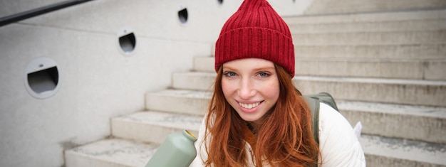 Gratis foto een glimlachende reiziger, een roodharige meisjetoerist, zit op de trap met een kolf en drinkt hete koffie uit een thermosfles terwijl