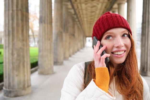 Gratis foto een glimlachende, schattige roodharige vrouw pleegt een telefoontje, houdt de telefoon in de buurt van het jaar en heeft een mobiel gesprek